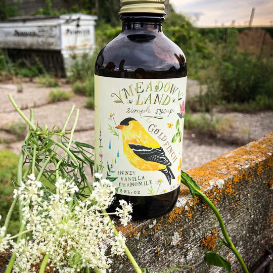 Bottle of Meadowland Honey Vanilla Chamomile syrup with a goldfinch label, surrounded by plants and flowers.