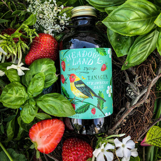 Bottle of Meadowland Tanager Strawberry and Basil Simple Syrup surrounded by strawberries and basil leaves